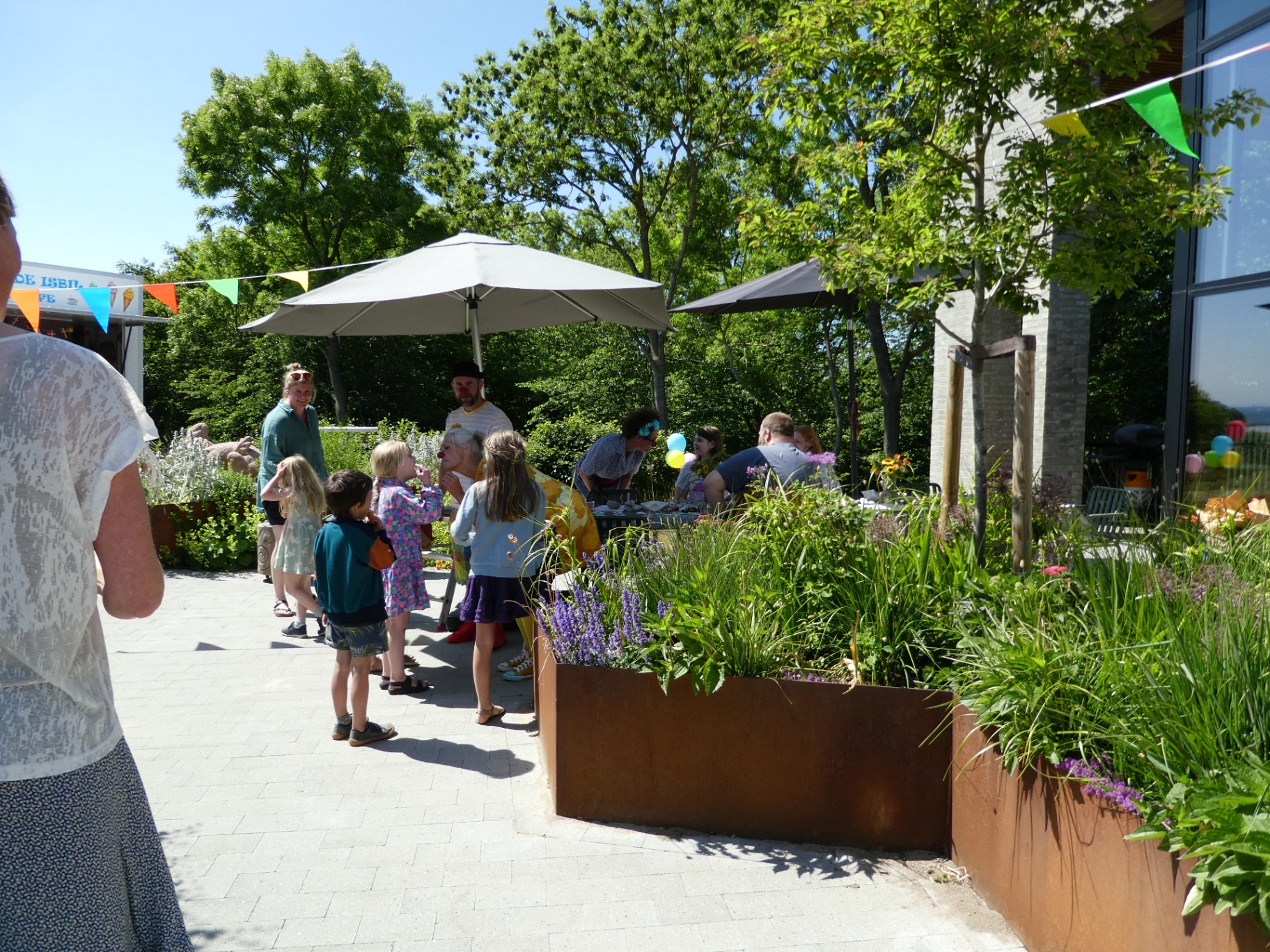 Sommerfest på terrassen — børn, hospitalsklovne og familier samlet i solen mellem blomsterbede